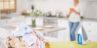 Woman in the kitchen with a ironing board and pile of laundry.