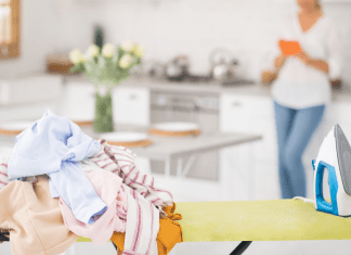 Woman in the kitchen with a ironing board and pile of laundry.