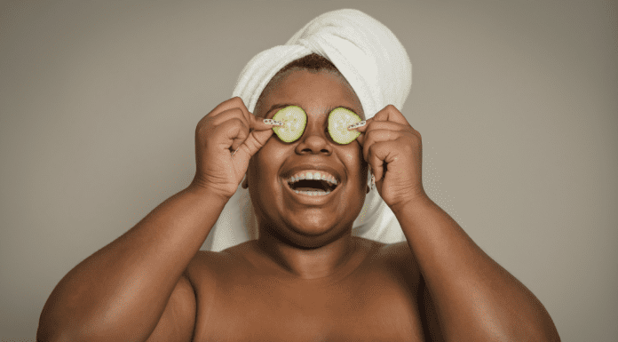 A woman with a towel on her head holding cucumbers over her eyes.