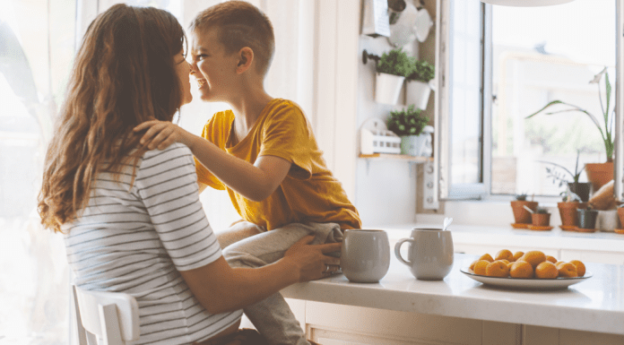 A boy sitting on the kitchen table hugging his mother.