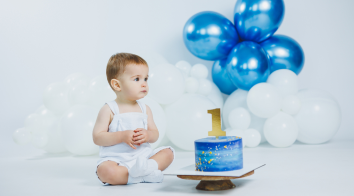 A boy sitting next to his first birthday cake and balloons.