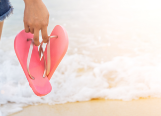 A woman holding flip flops by the ocean.