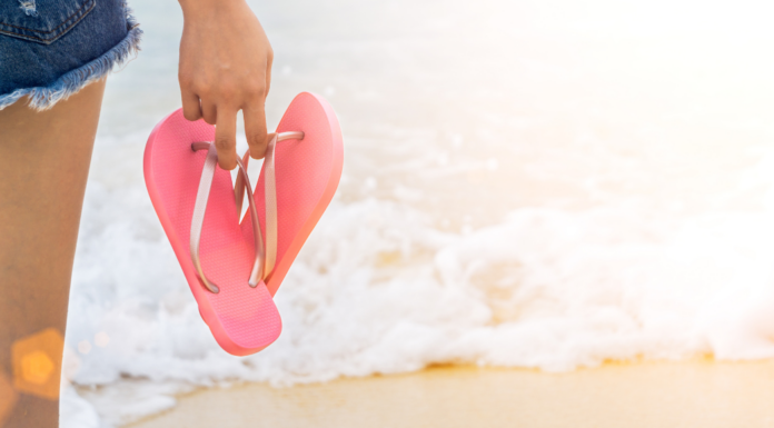 A woman holding flip flops by the ocean.