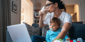 A busy mom drinking coffee, holding her son, and working on the computer.