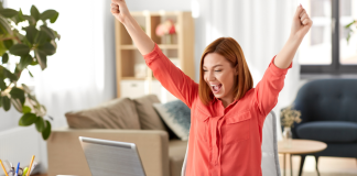 A woman raising her hands in joy as she sits at the computer.