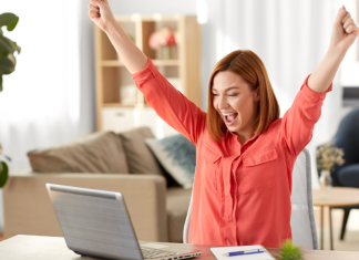 A woman raising her hands in joy as she sits at the computer.