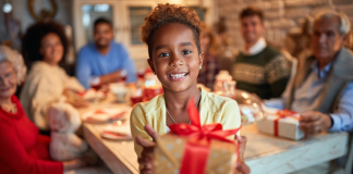 A girl holding a gift in front a family celebrating at the table.