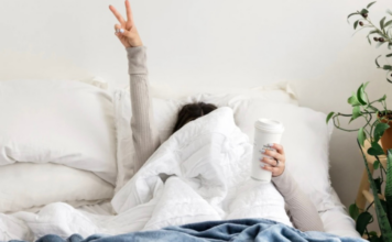 A woman hiding under the covers making a peace sign and holding a cup of coffee.