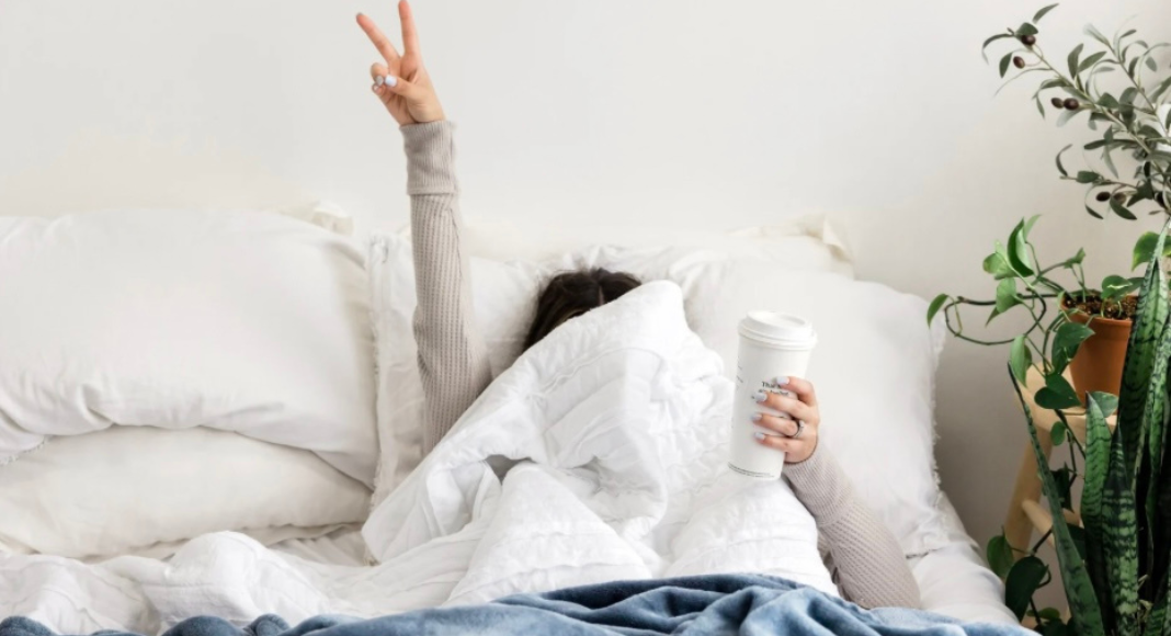 A woman hiding under the covers making a peace sign and holding a cup of coffee. 