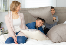 A mother in bed with her boys hiding behind pillows.
