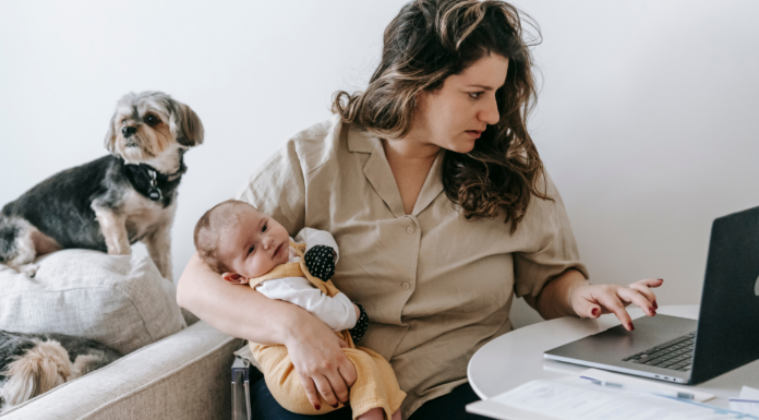 A woman working while holding her baby and the dog sits behind her.