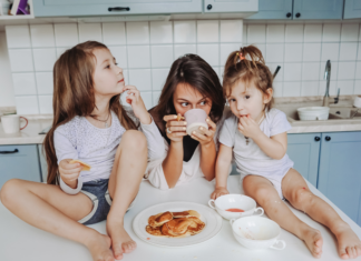 I Live For My Kids A mom drinking coffee while girls eat pancakes on the counter.