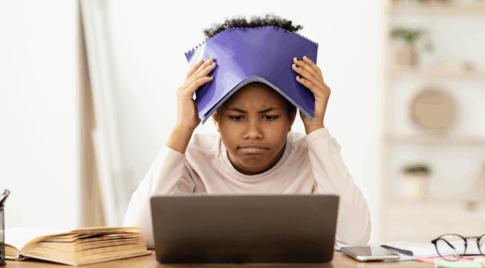 A girl holding a notebook over her head while looking at a computer.
