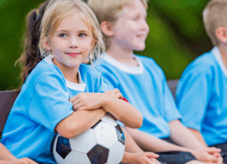 Are Youth Sports Losing Their Innocence? A girl holding a soccer ball while kids sit on a team bench.