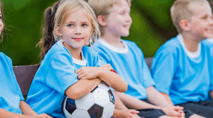 A girl holding a soccer ball while kids sit on a team bench.