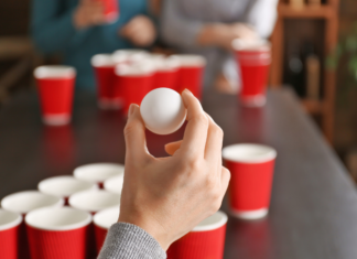 A hand holding a ping pong ball in front of a beer pong table with red solo cups.