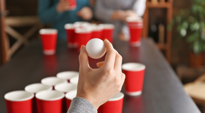A hand holding a ping pong ball in front of a beer pong table with red solo cups.