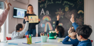 A teacher standing in front of children raising their hands.