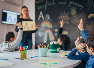 A teacher standing in front of children raising their hands.
