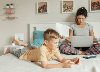 A woman working on her computer with her son laying on her bed.